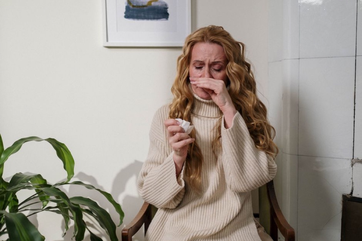Woman indoors using a tissue to relieve allergy symptoms, houseplant visible in background